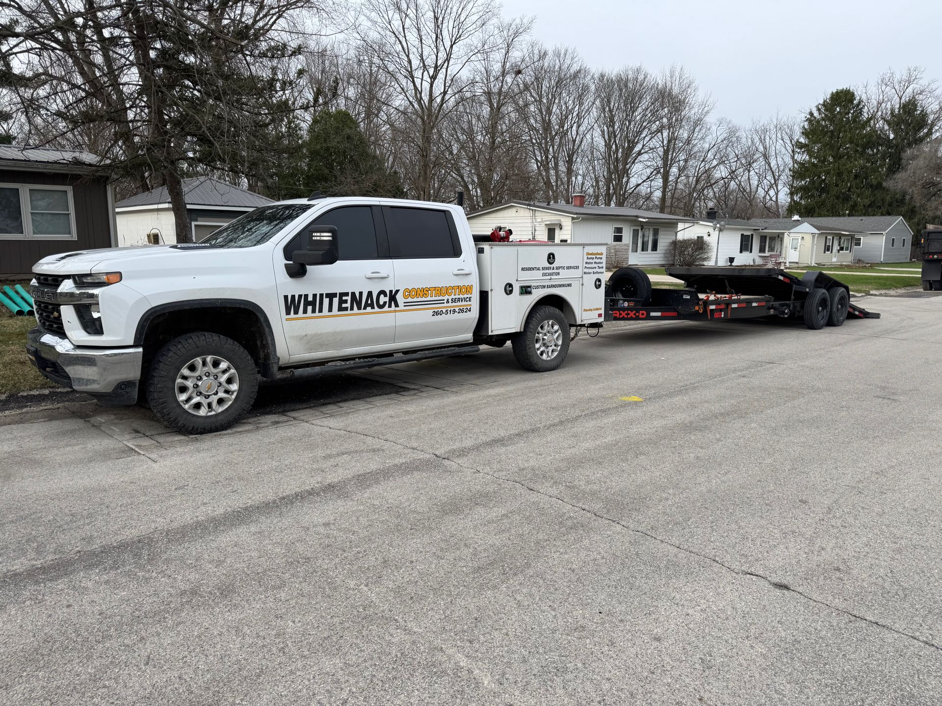 White utility truck towing a flatbed trailer parked on a residential street under cloudy skies