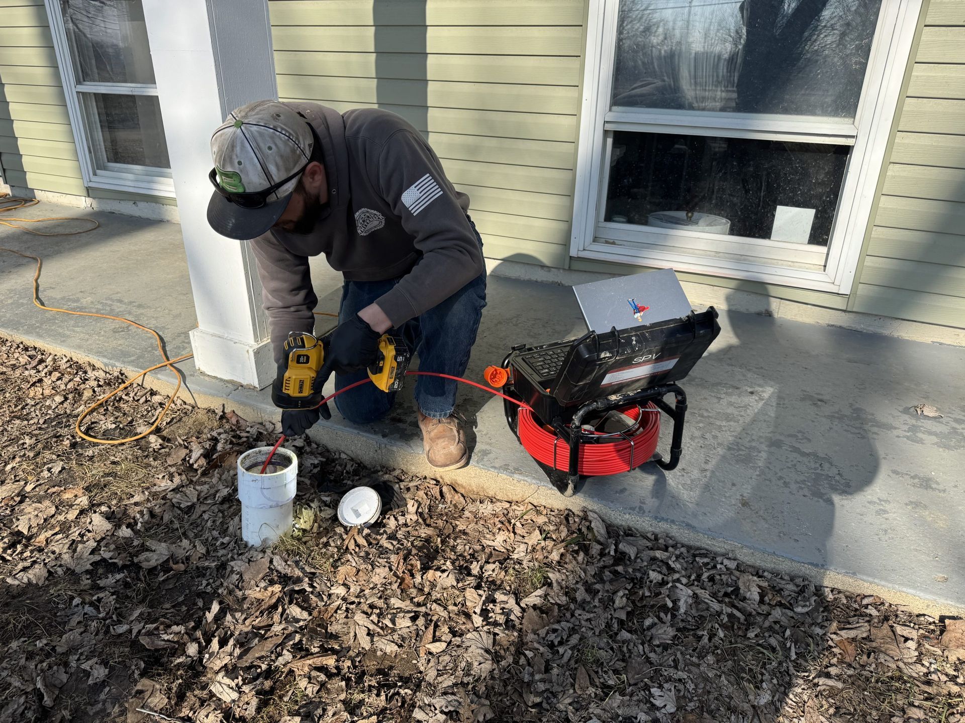 Worker drilling concrete beside a house, with tools and a red blower on a leaf-covered walkway