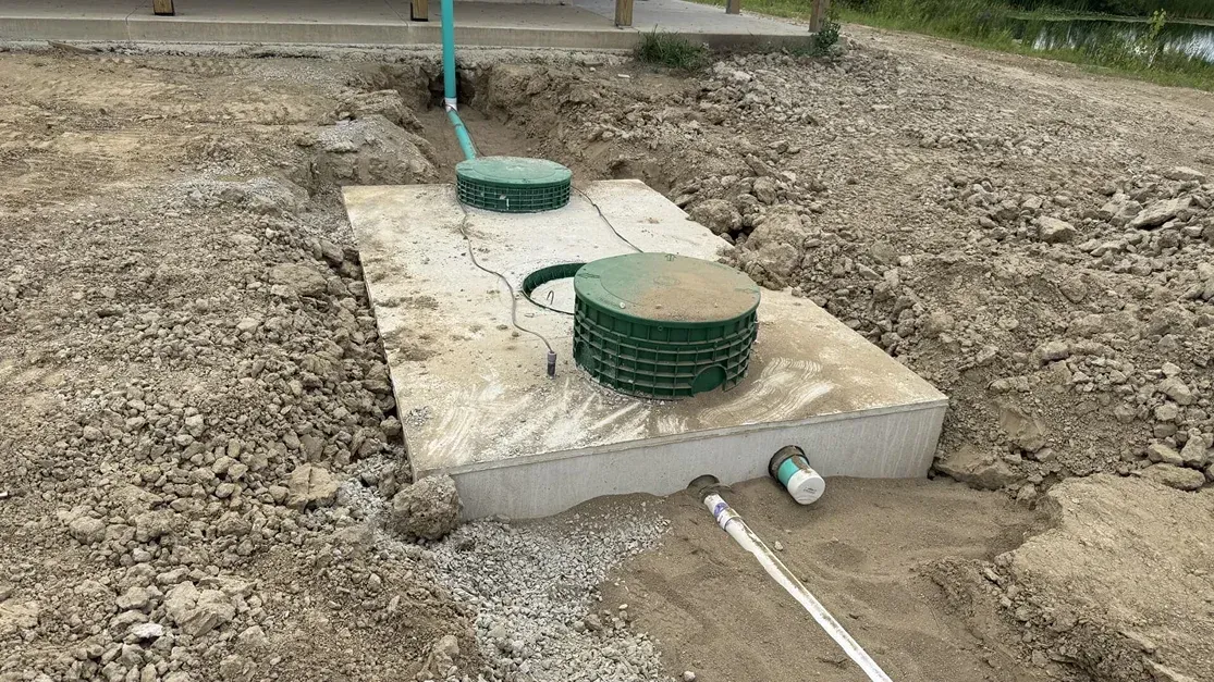Concrete utility boxes with green covers on a gravel lot, with pipes and a hose nearby