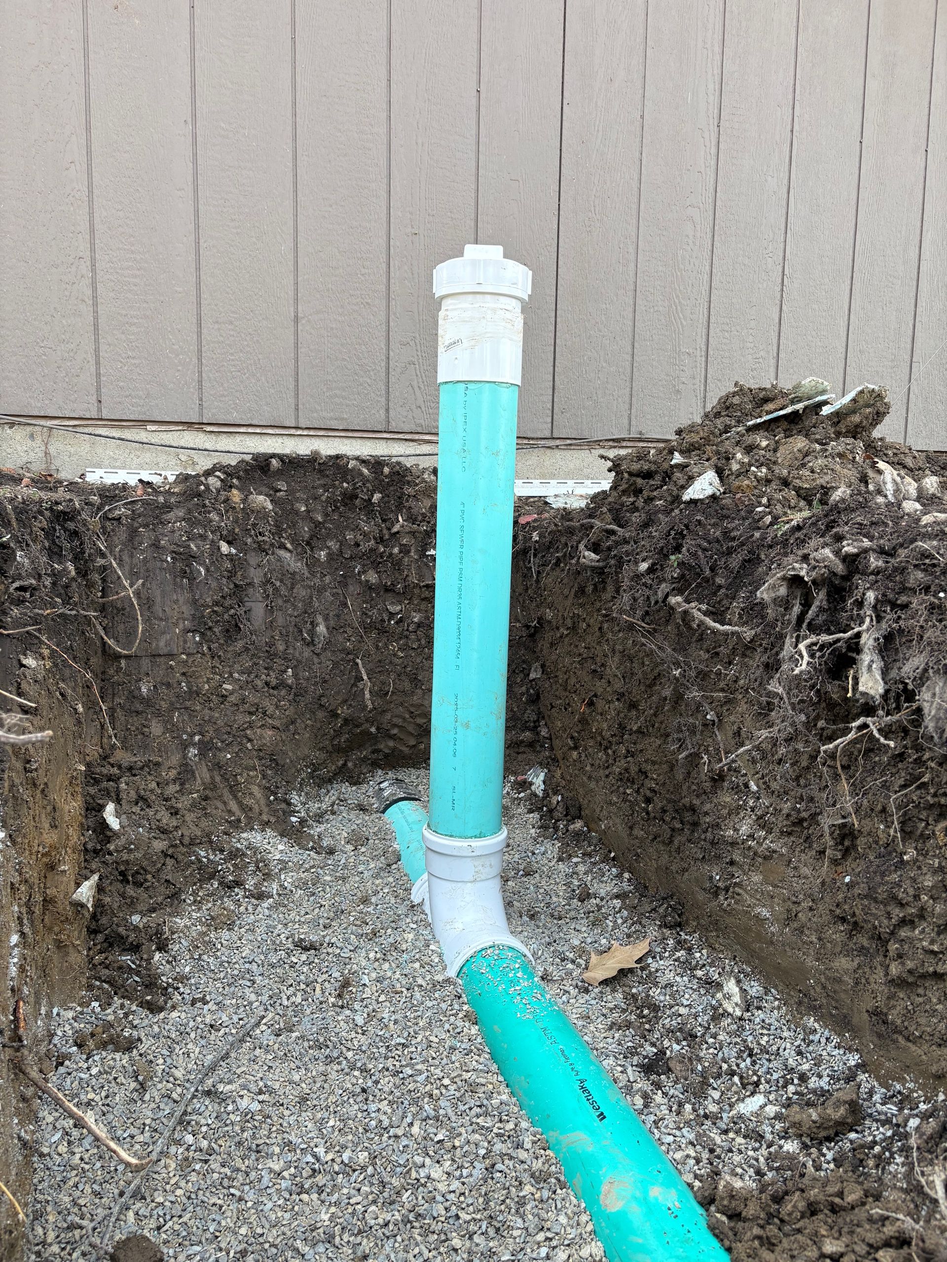 Exposed turquoise PVC pipe in a dirt trench beside a beige wall