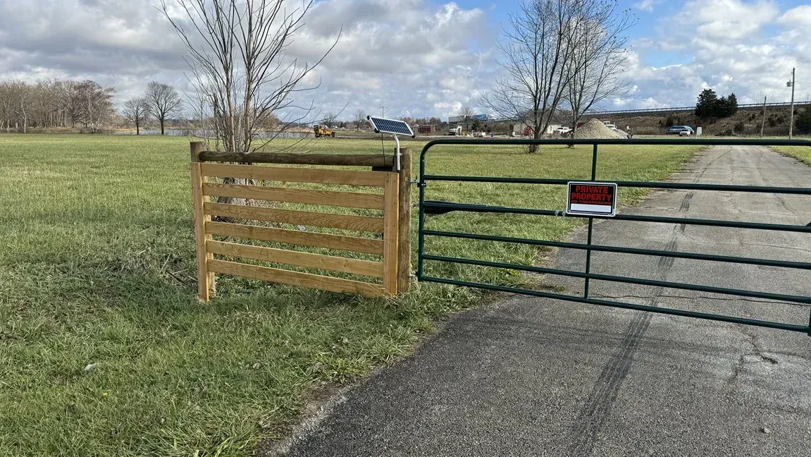 Wooden and metal gate across a gravel driveway beside an open grassy field under a cloudy sky