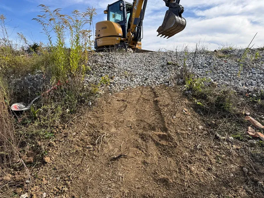 Excavator digging a dirt path beside a gravel mound under a clear blue sky.