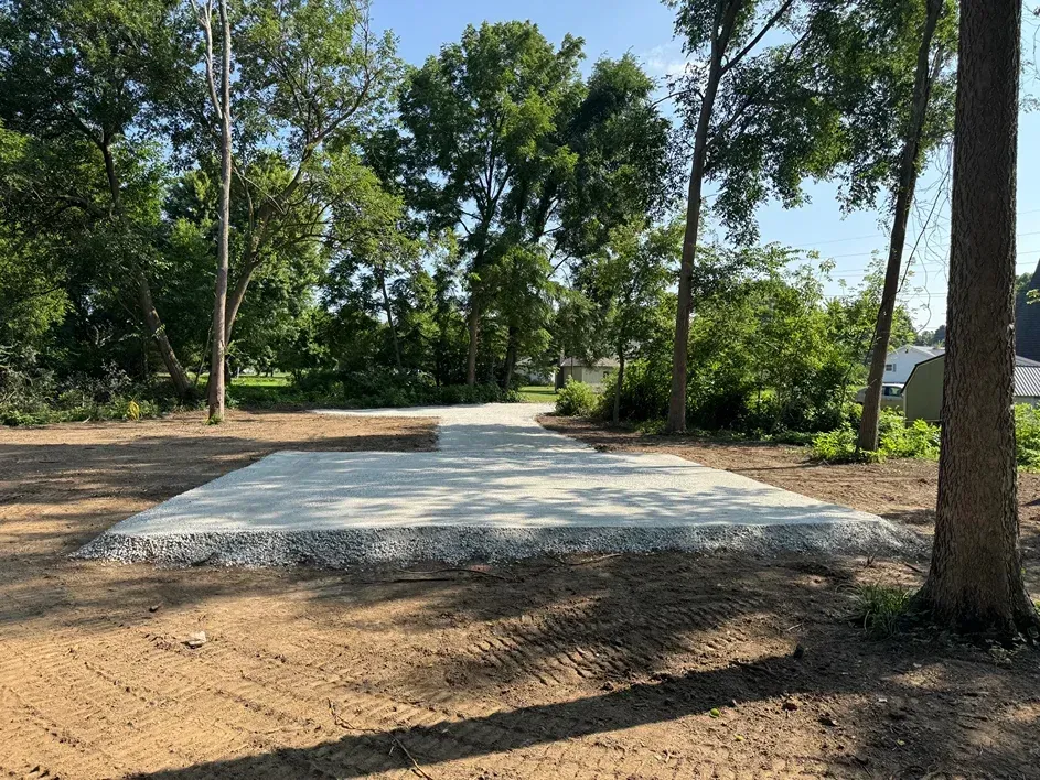 New concrete pad in a wooded park clearing, with trees and dirt around it.