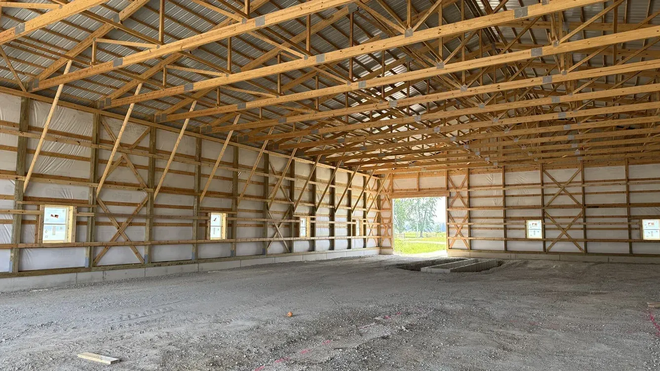 Empty wooden barn interior with exposed trusses, gravel floor, and sunlight through a central opening.