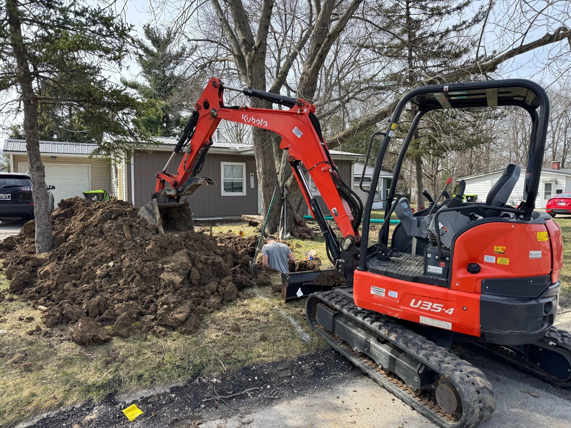 Red excavator dumping dirt beside a small house in a yard with trees.