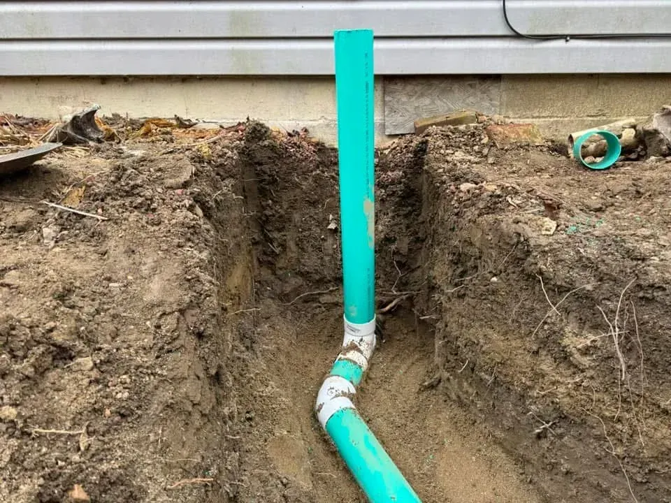 Freshly dug trench with a turquoise pipe and vertical standpipe beside a house foundation