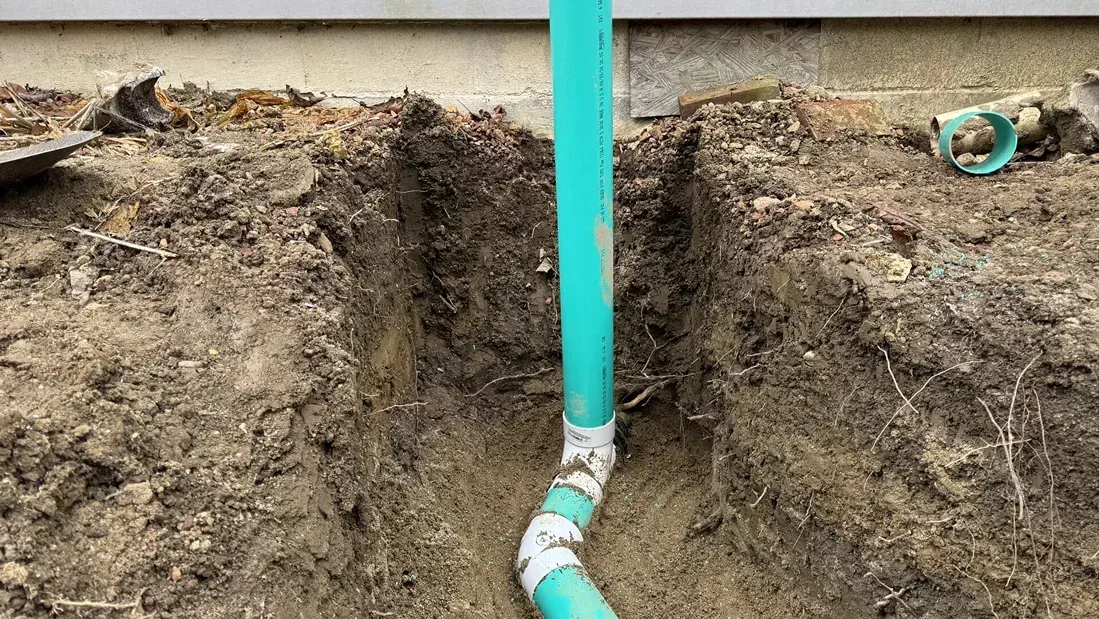 Exposed turquoise pipe in a dirt trench beside a building foundation