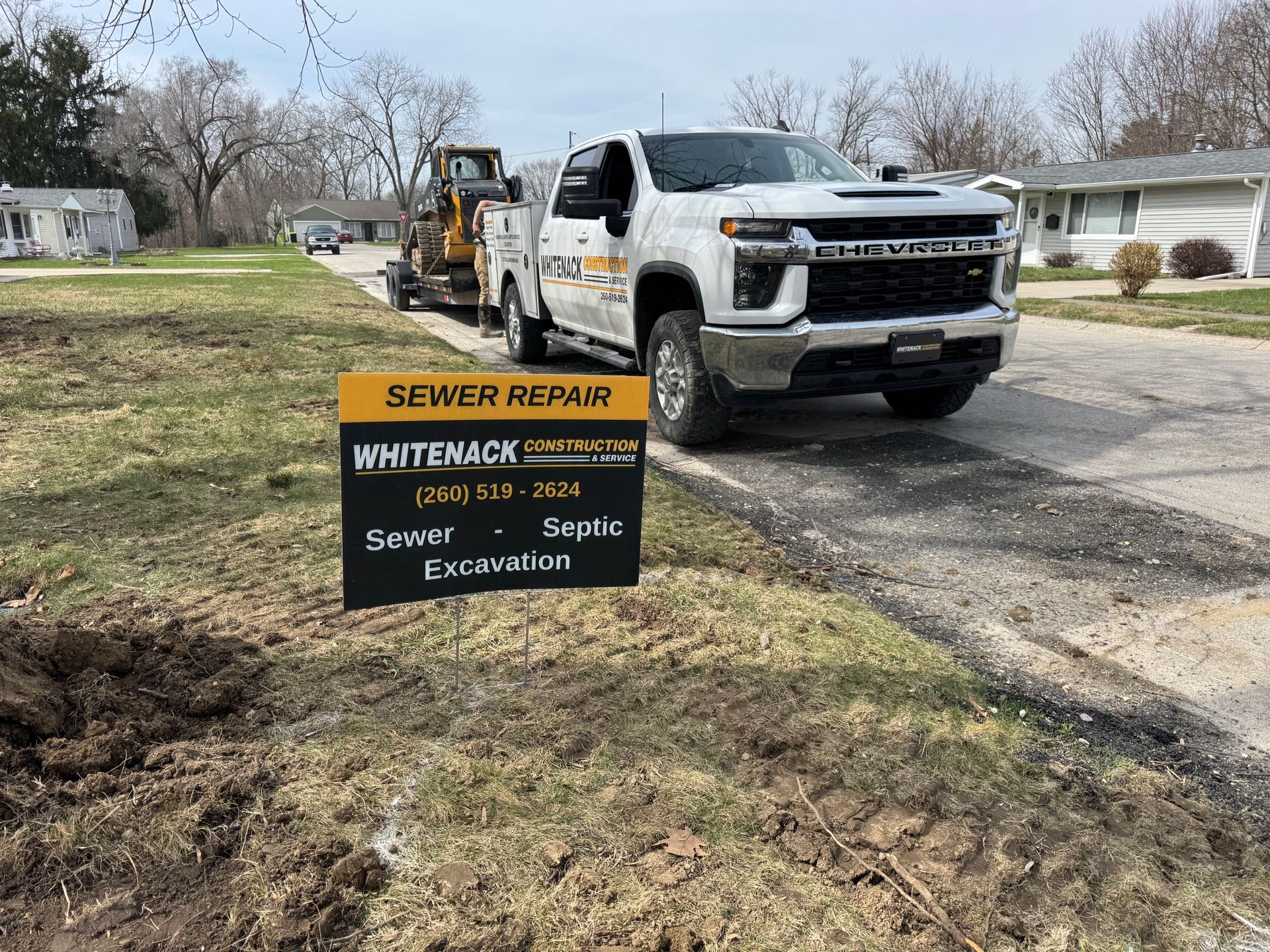 White utility truck beside a sewer repair sign on a suburban roadside, with a backhoe in the background.