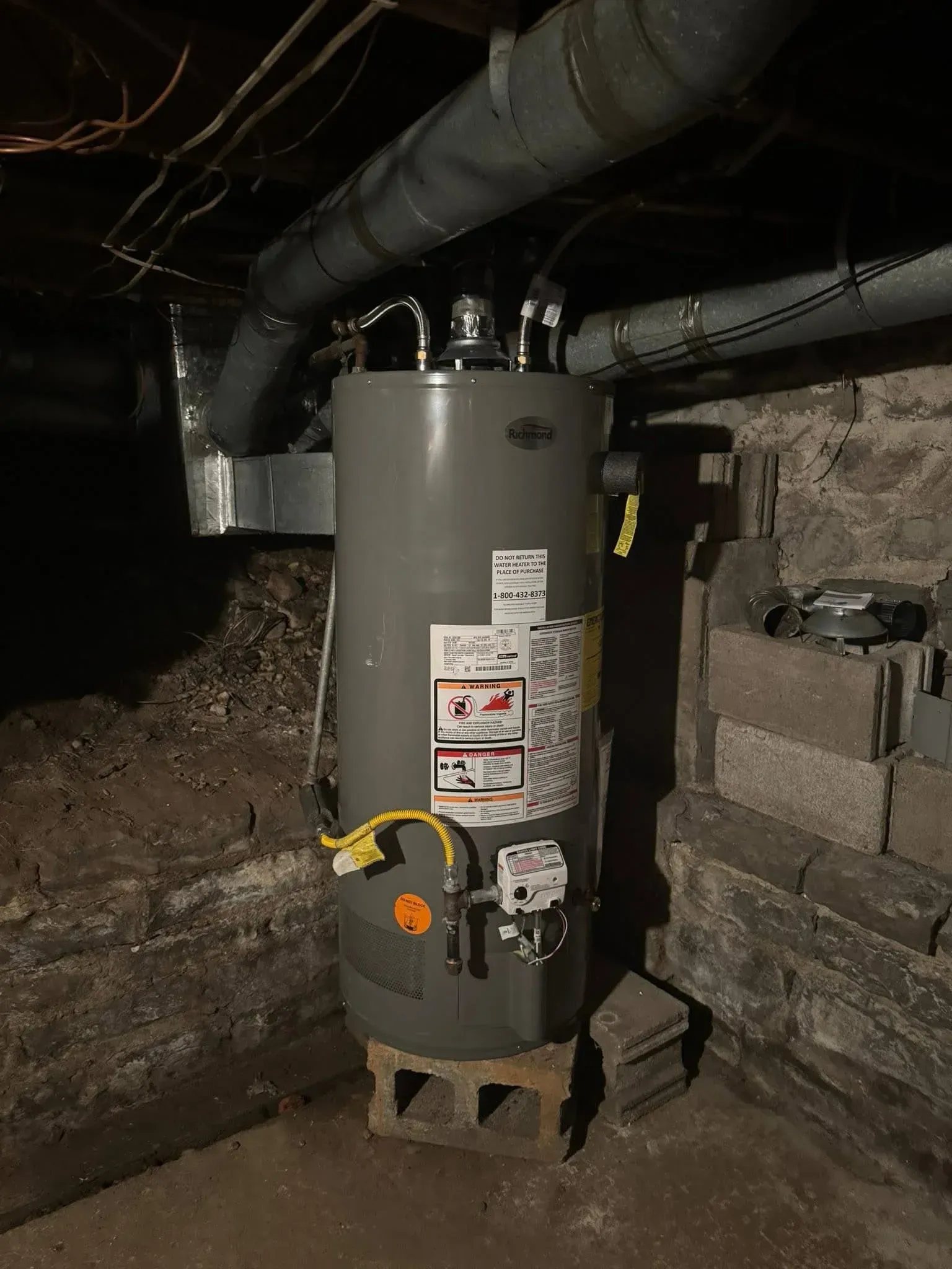 Gray water heater with warning labels and yellow valve handle in a basement, under exposed pipes.