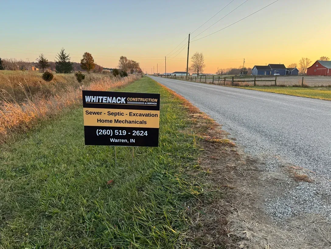 Roadside real estate sign on grassy verge beside a gravel road at sunset