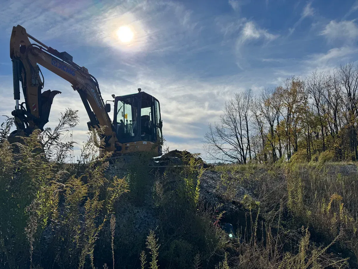 Excavator on a grassy hillside under a bright sun, with trees and blue sky in the background
