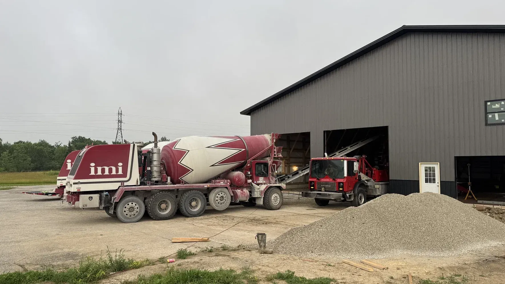 Red-and-white cement mixer trucks parked beside a large metal barn on a gravel lot.