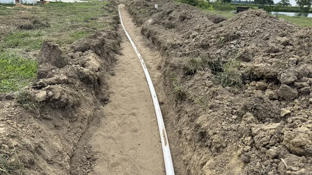 A white pipe runs through a trench in muddy ground beside patches of grass.