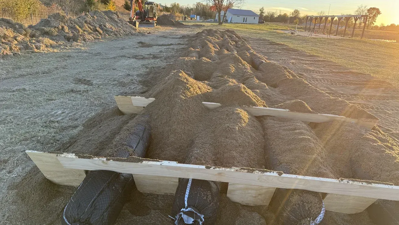 Gravel road construction with white barriers and dirt mounds at sunset, viewed from a vehicle