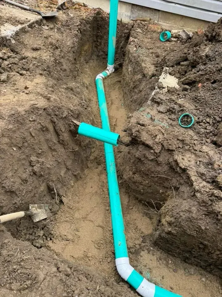 Bright blue PVC pipe installed in a dirt trench beside a building foundation.