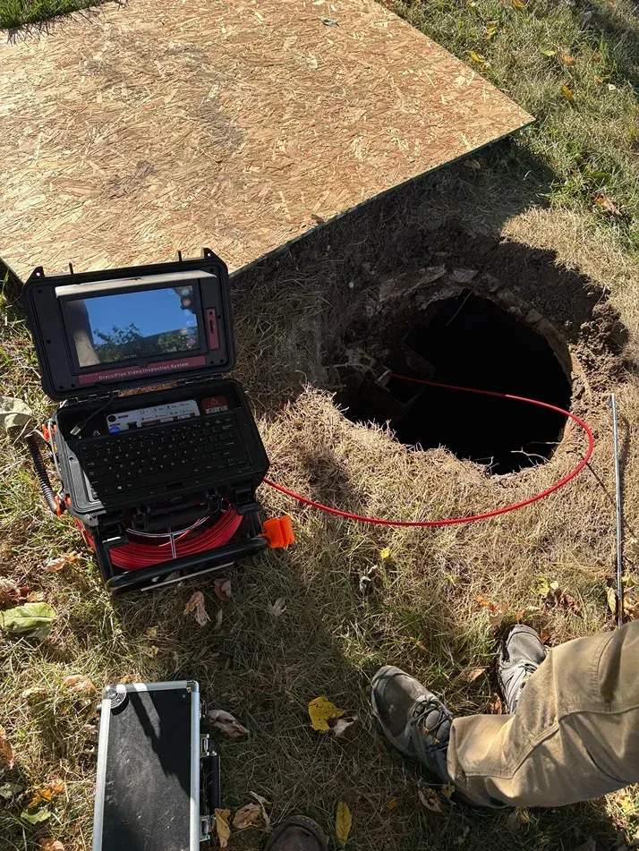 Laptop beside open manhole with red cable, worker’s boots nearby, and a small utility trailer on grass