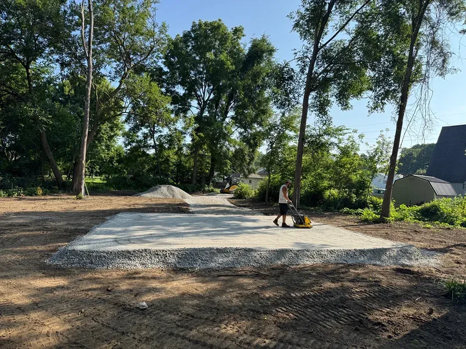 Person working on a gravel pad in a wooded clearing with trees and a small building nearby