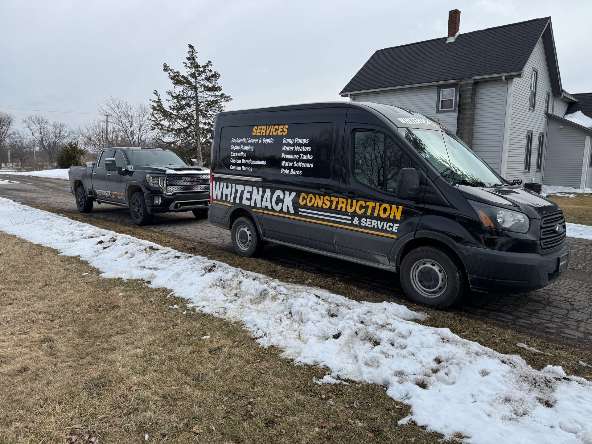 Black construction van parked by a snowy roadside beside a house and pickup truck