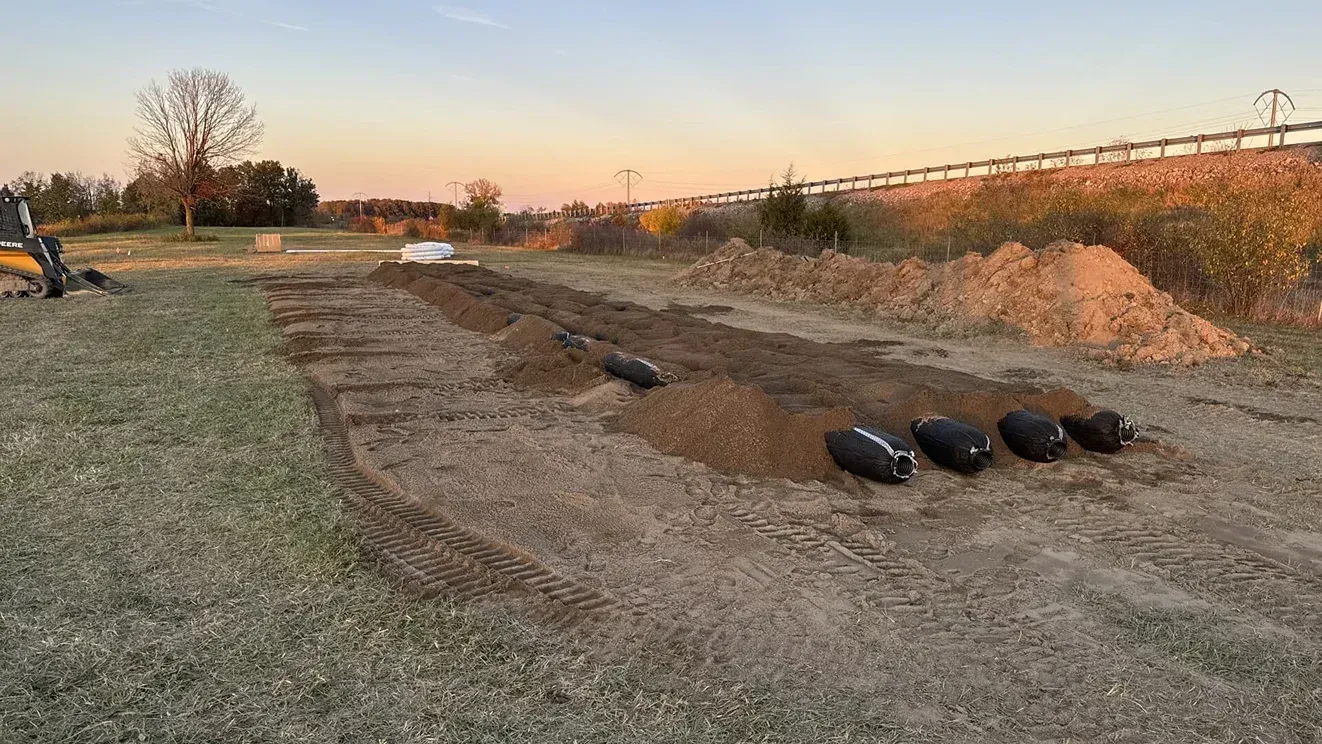 Grassy construction site with a dirt trench, black pipes, and an orange sunset sky