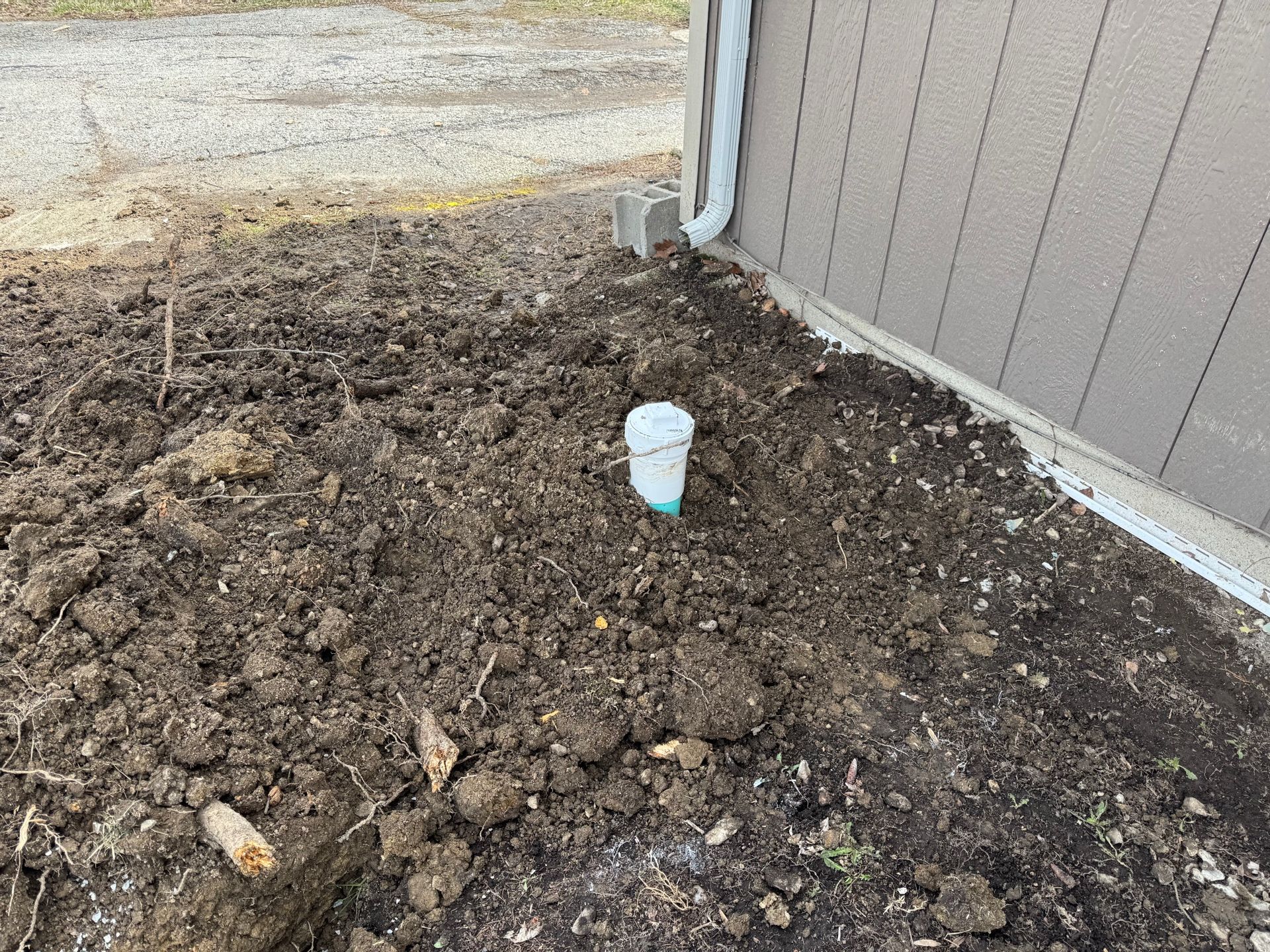 Dirt pile beside a house wall with a white cup stuck in the soil near a driveway.