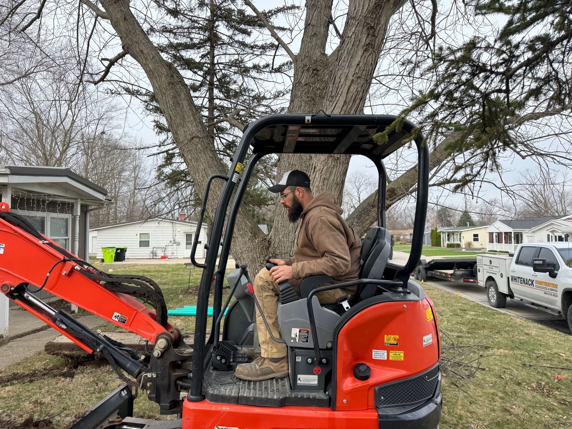 Man sitting on a compact orange excavator under leafless trees in a yard