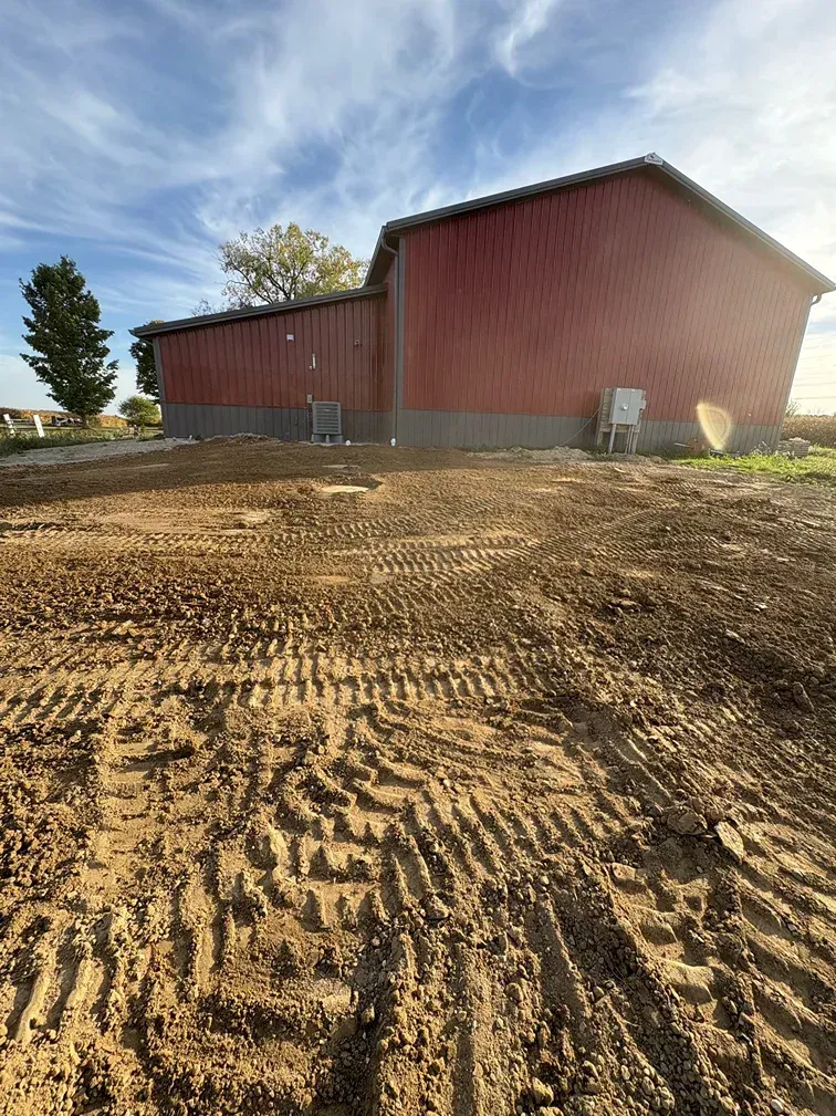 Red barn-like building on a freshly graded dirt lot under a partly cloudy sky.
