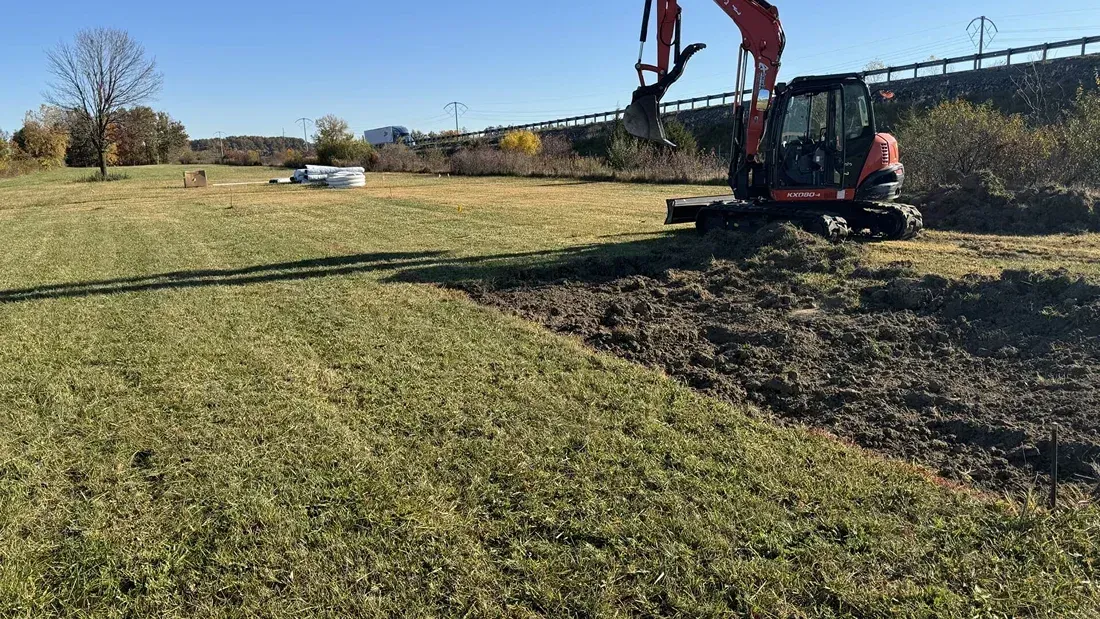 Excavator digging a trench beside a grassy field on a sunny day