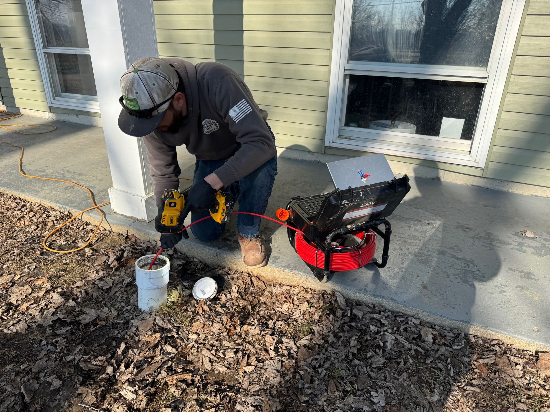 Worker kneeling on a porch, using a drill near a bucket and tool bag beside a house wall.