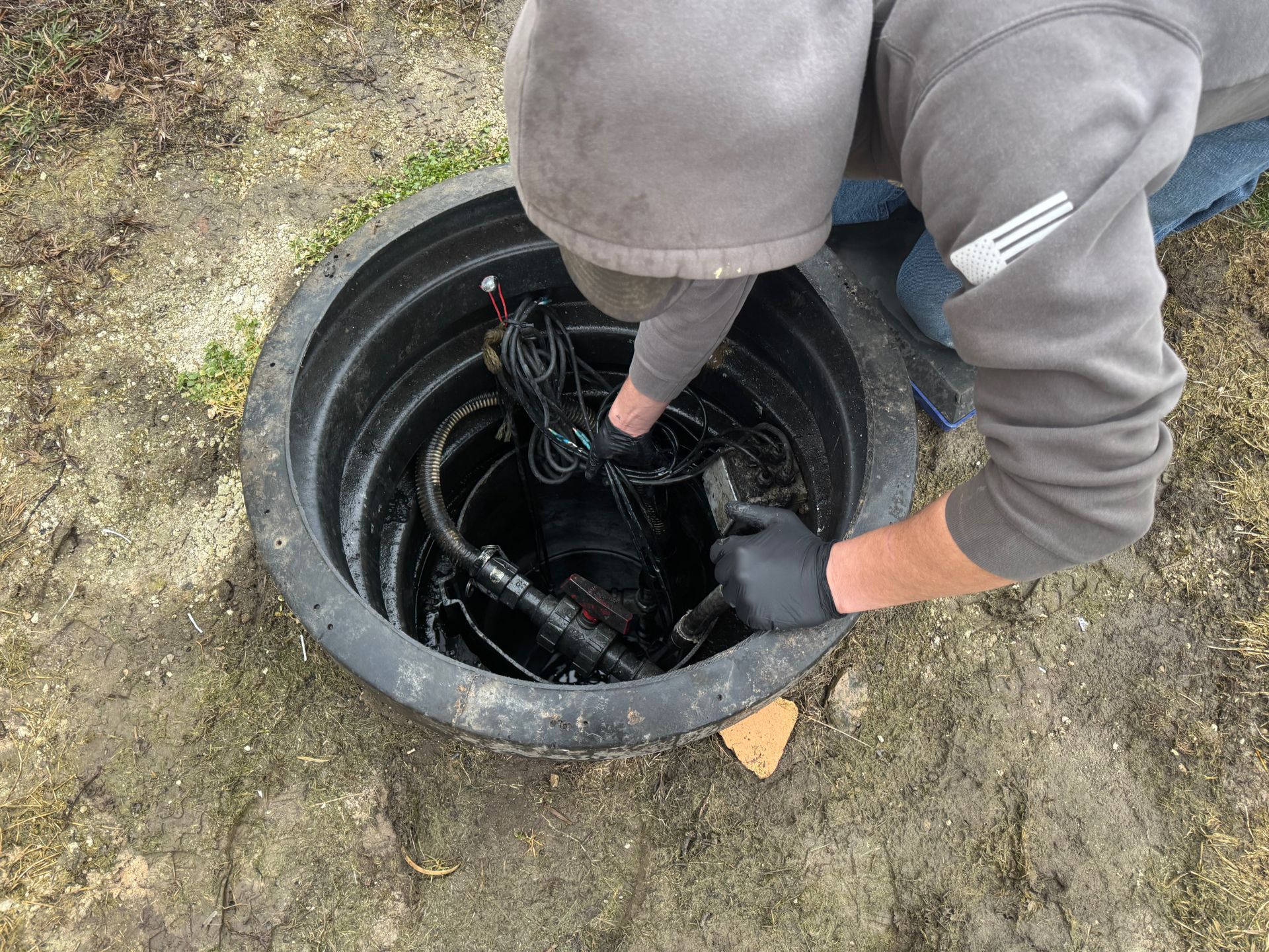 Person in gray hoodie reaching into an open manhole with exposed cables.