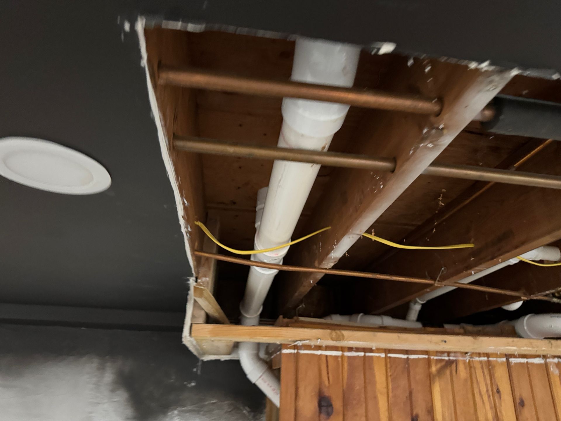 Exposed ceiling framing with white ductwork, pipes, and yellow electrical wires beside a wooden wall panel.