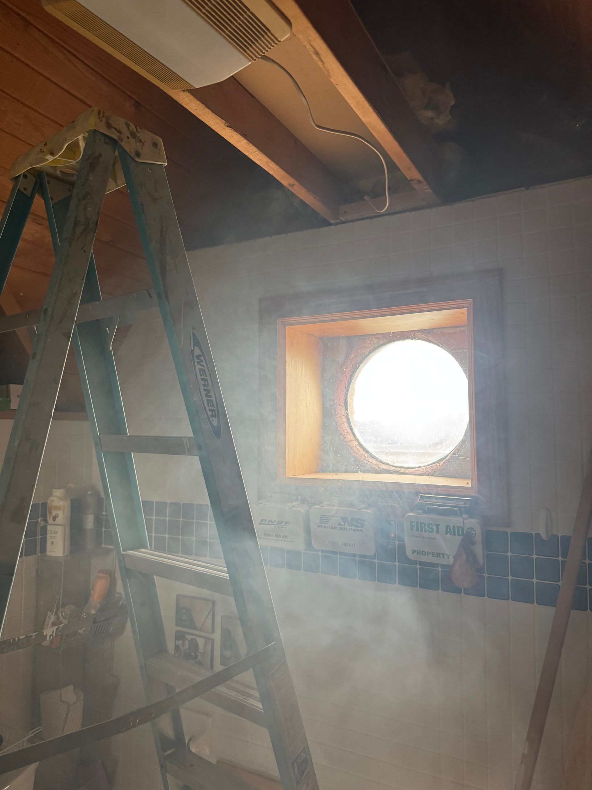 Indoor room under renovation with ladder, exposed ceiling beams, and a small round window letting in bright light