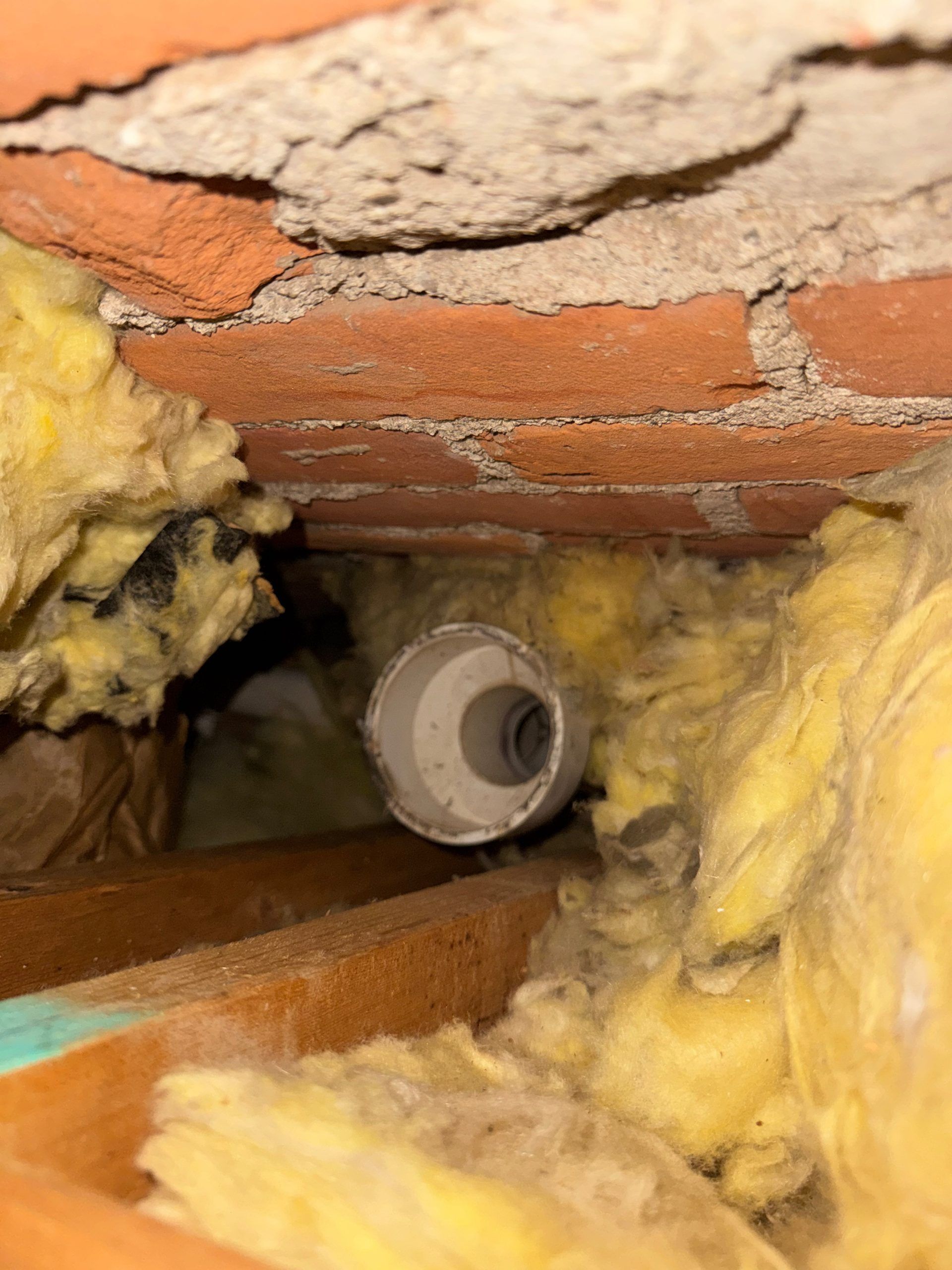 Yellow insulation around a gray PVC pipe inside a wall cavity with exposed brick and wood framing