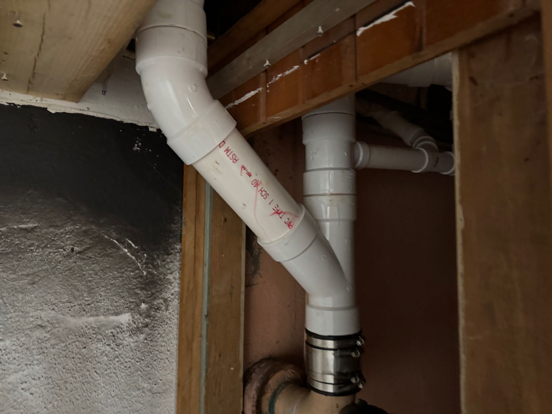 White PVC pipes and a metal clamp under a wooden ceiling, next to a black wall panel.