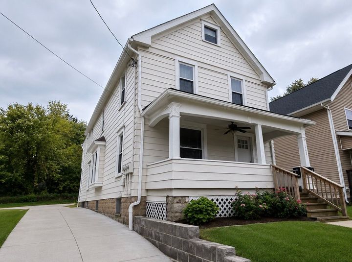 White two-story house with a front porch, green lawn, and a stone retaining wall along the sidewalk.