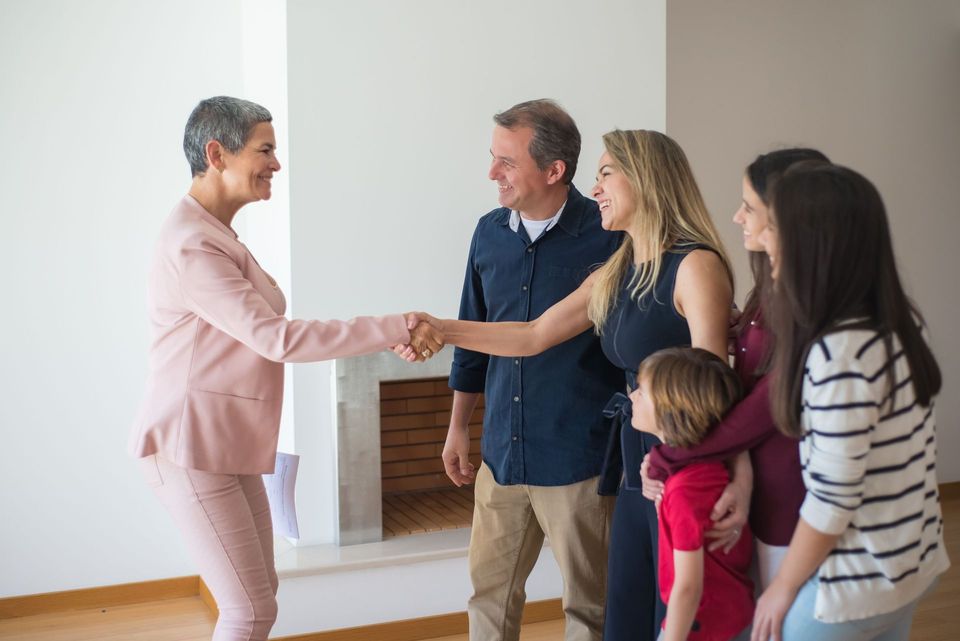 Two people shaking hands in a bright living room while others look on with a moving box nearby