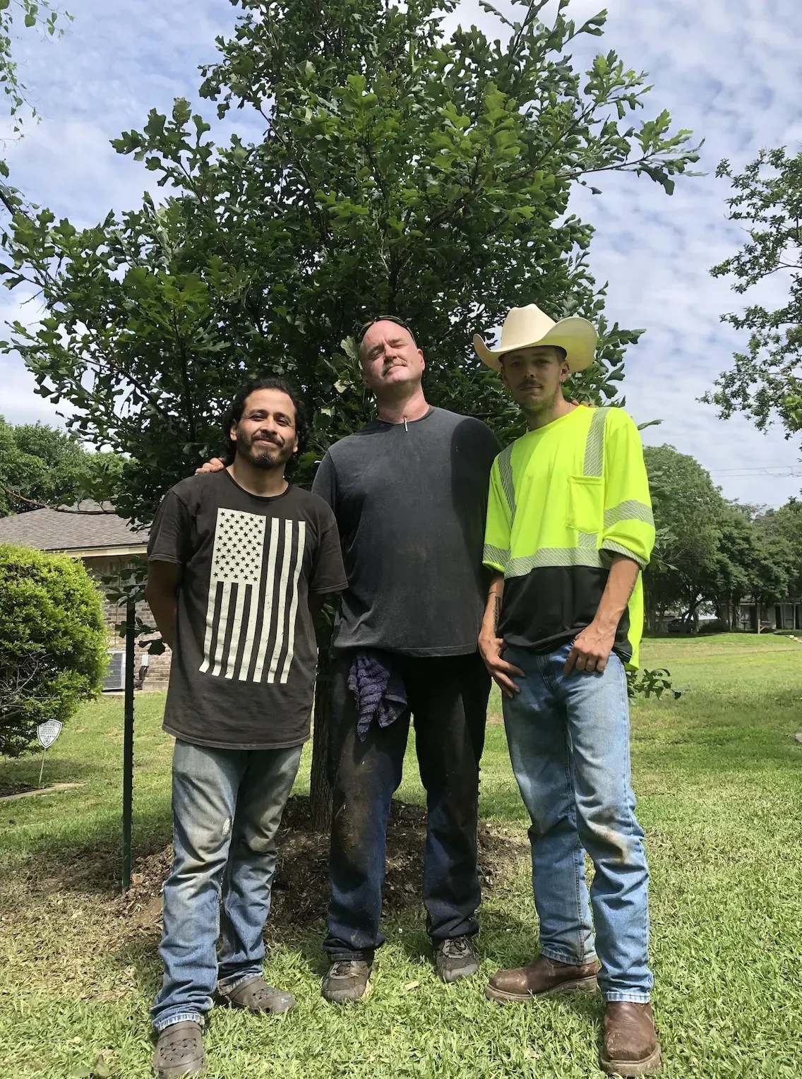 Three people standing together in a grassy park, with trees and a house in the background.