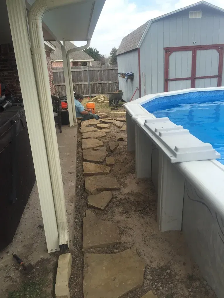 Narrow stone walkway beside a house and above-ground pool, leading past a shed in a backyard.