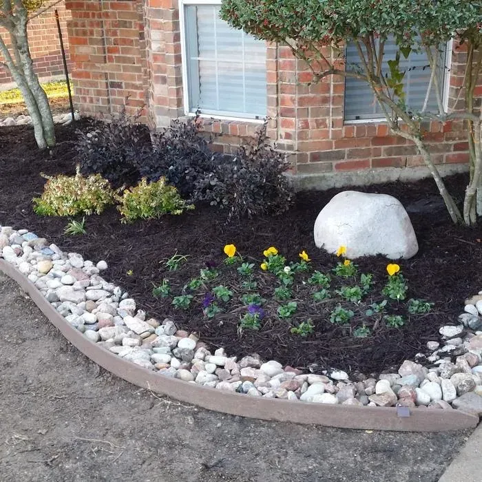 Front yard flower bed with yellow blooms, white stones, mulch, and a large decorative rock beside a brick house