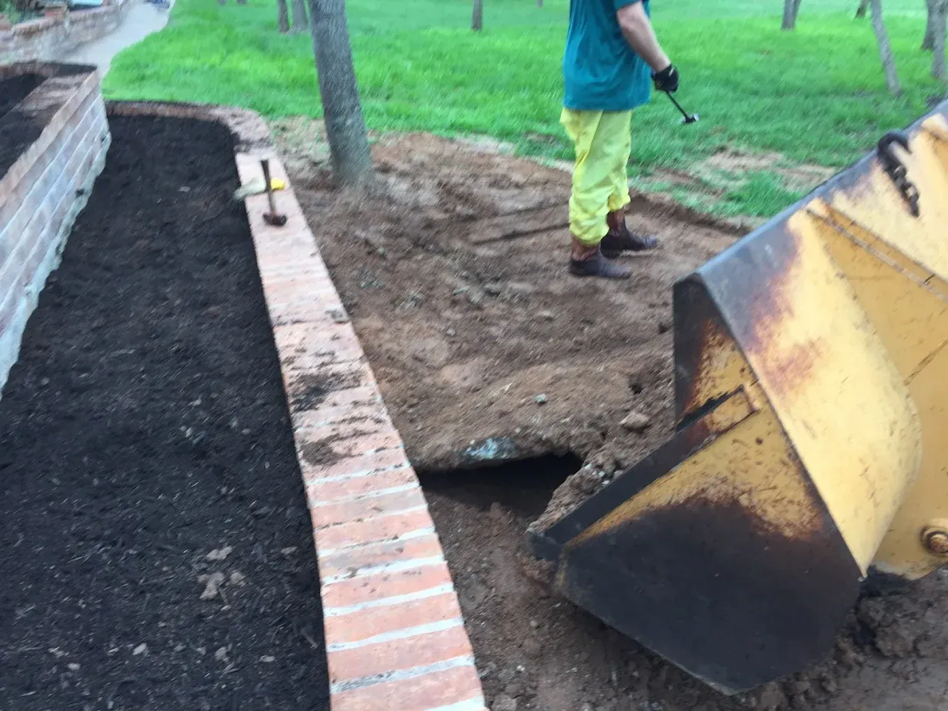 Worker in a green shirt stands beside a loader bucket dumping dirt next to a brick walkway.