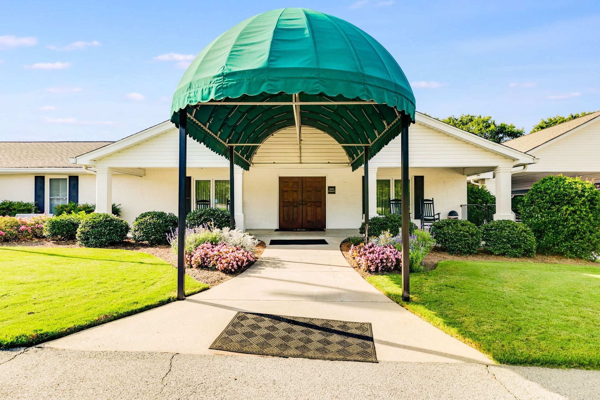 Covered entrance to a white building with a green awning, walkway, and landscaped lawn