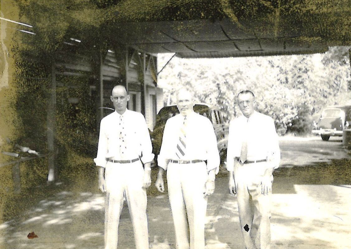 Three people stand under a shaded structure on a sunlit road, with trees and a parked car in the background.