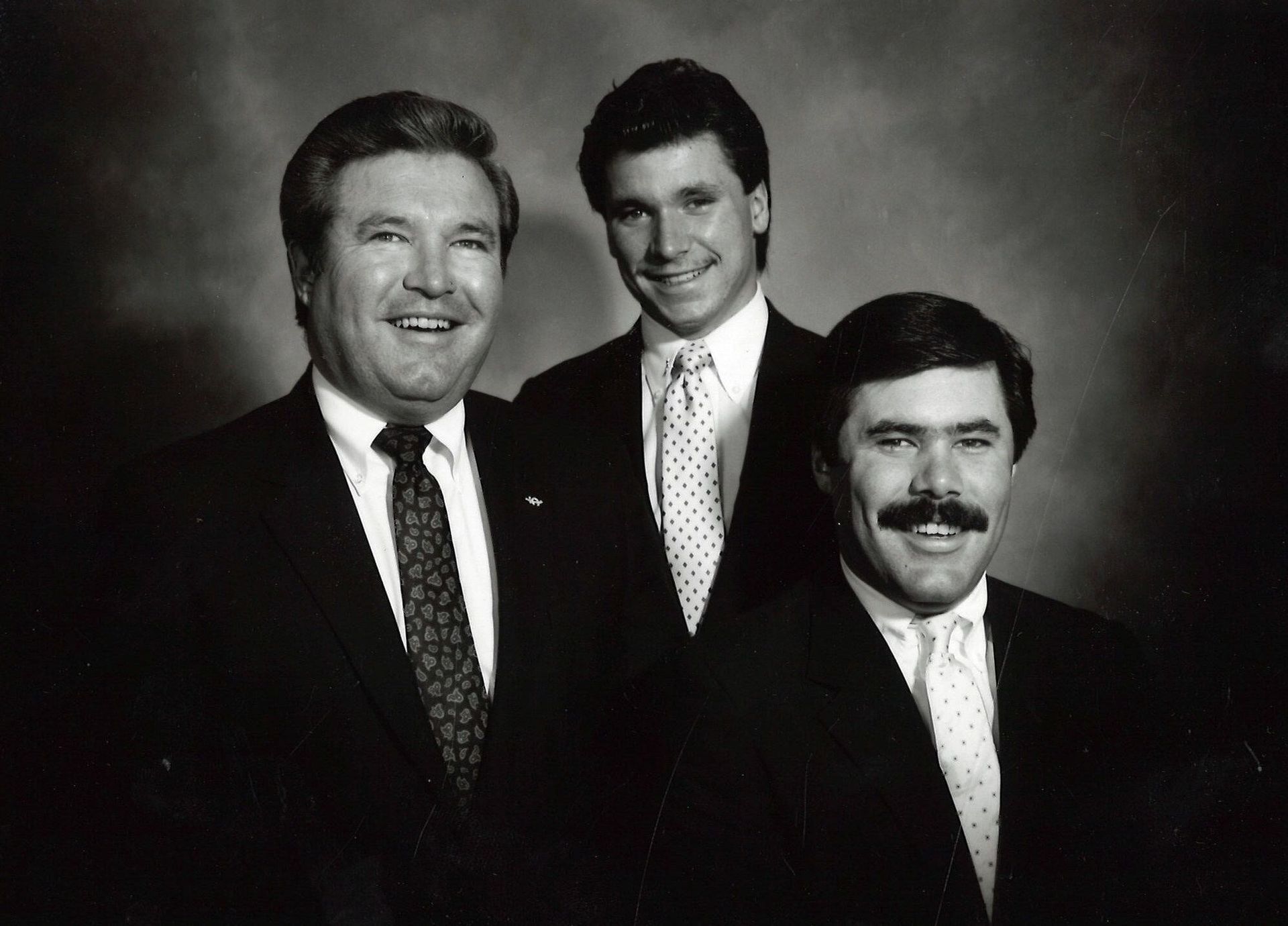 Three men in suits posing for a formal portrait, two standing behind one seated in front.