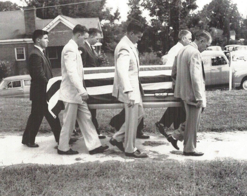 Five people in suits carrying a casket outdoors, walking past parked cars and a house.