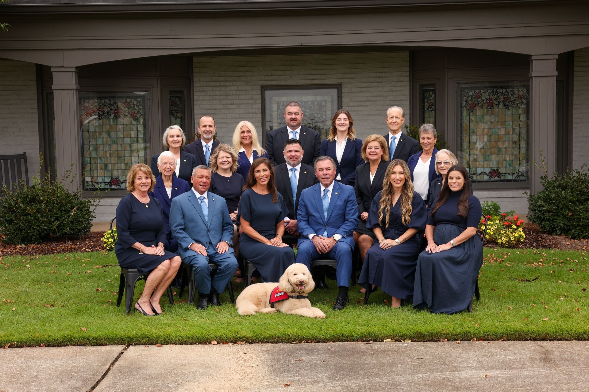 Formal group portrait of 16 people and a dog posed in front of a house