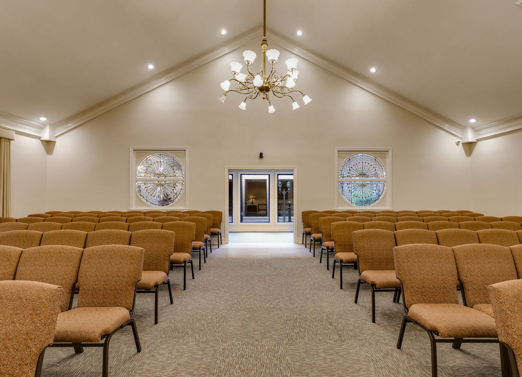Empty ceremony hall with rows of tan chairs, chandeliers, and a central aisle leading to double doors.
