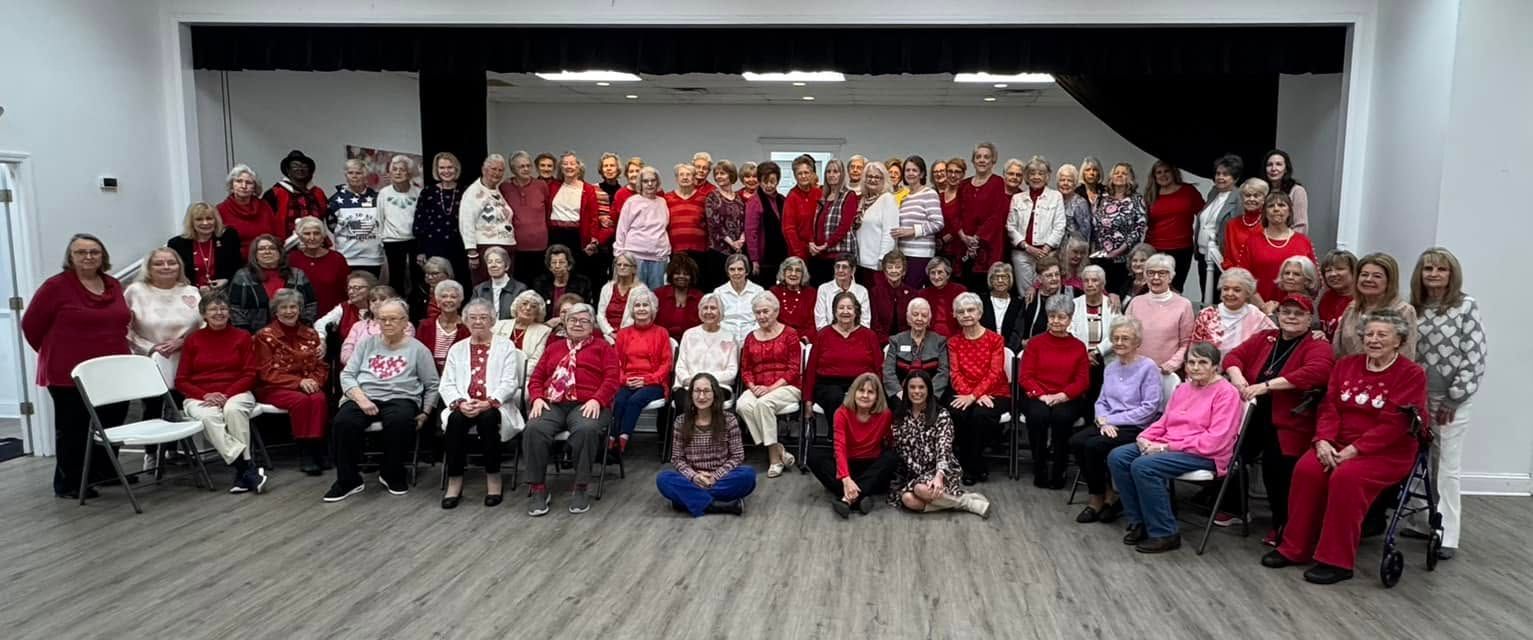 Large group posing in a hall, mostly wearing red and white, with a black-and-white backdrop and one blue bag in front