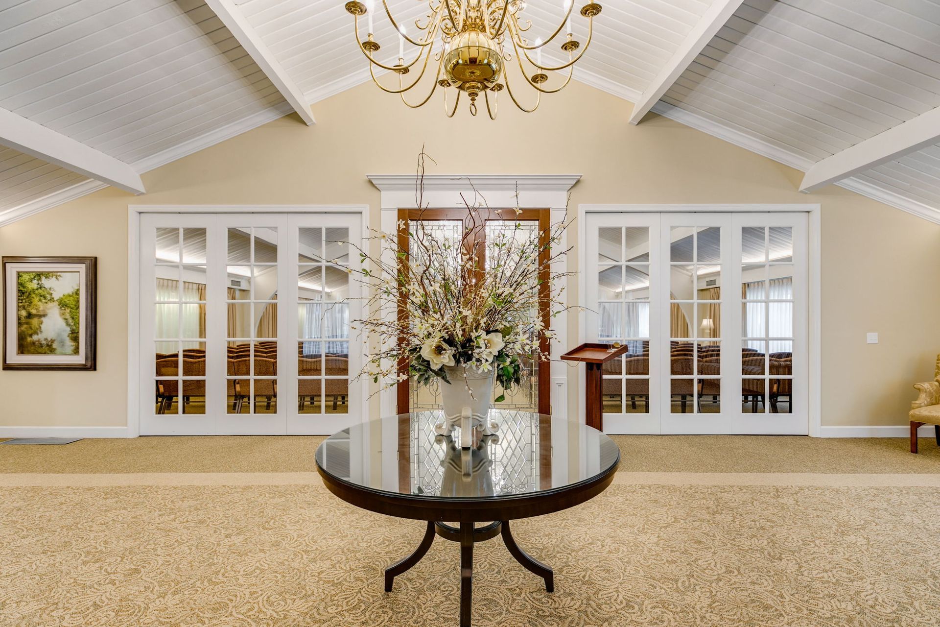 Spacious lobby with chandeliers, glass doors, a round table with flowers, and a podium on carpeted floor.