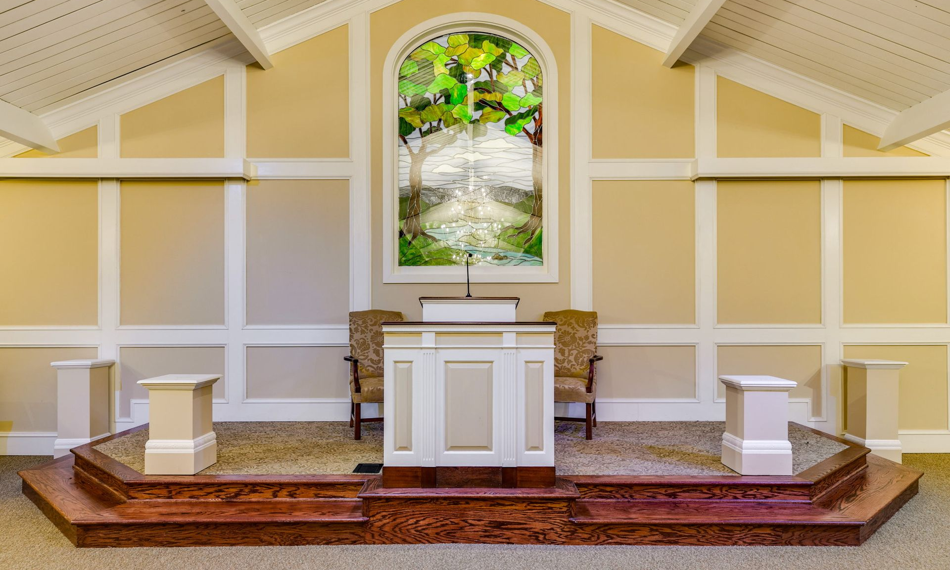 Bright church altar with white paneling, red carpet, and stained-glass window above the pulpit