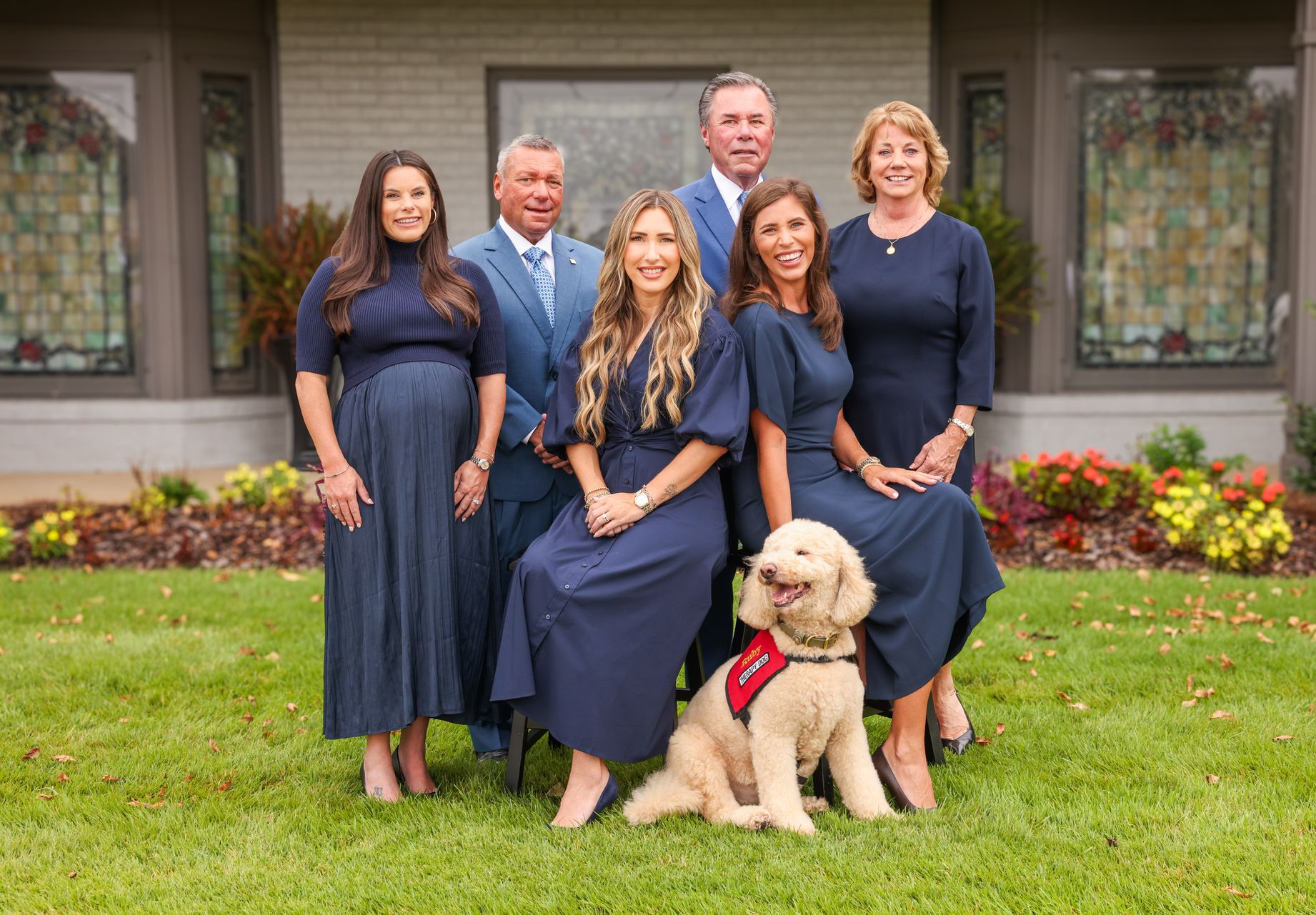 Six people pose on a lawn before a house, with a cream-colored dog wearing a red bandana in front.