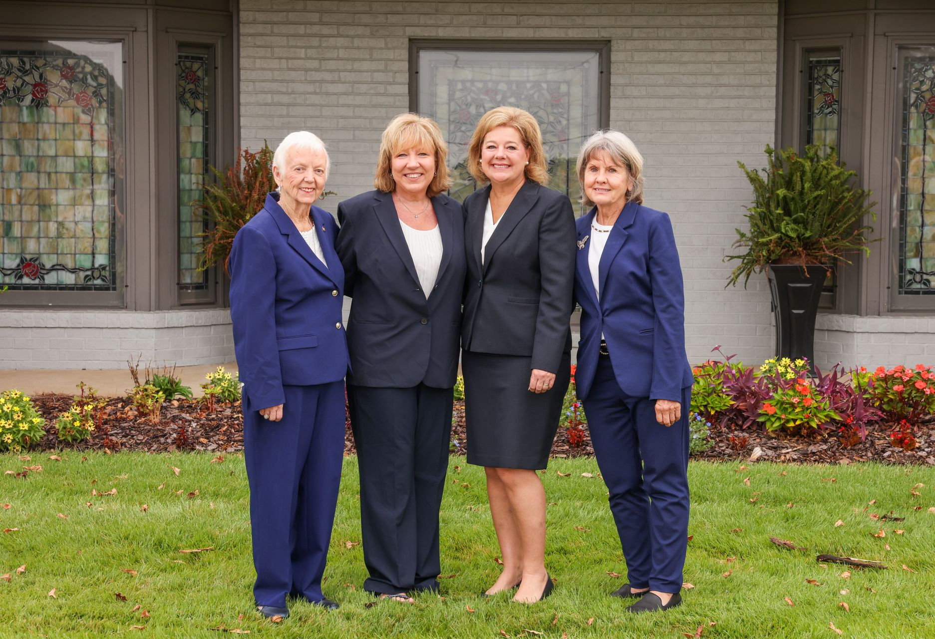 Four women in navy suits standing together outside a building with flowers and a lawn.